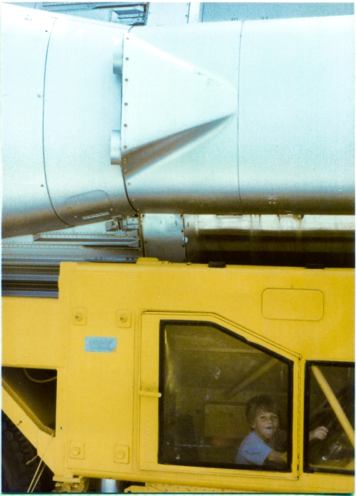 Kai MacLaren sits, enjoying himself hugely, hands on the steering wheel, in the “RIGHT TILLERMAN ENCLOSURE” of the “Guided Missile Semitrailer” which is used to transport the Atlas ICBM, a small part of which can be seen above him, at the Air Force Space and Missile Museum, Cape Canaveral, Florida. Click the links in the text box at left for more information on the Atlas missile and its transport semitrailer.  Photograph by James MacLaren.